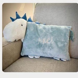 May include: A plush dinosaur-shaped pillow in white and light blue, with the name "Noel" embroidered on the pillow. The dinosaur has blue spikes and a fluffy white head. The pillow is resting on a gray upholstered chair.