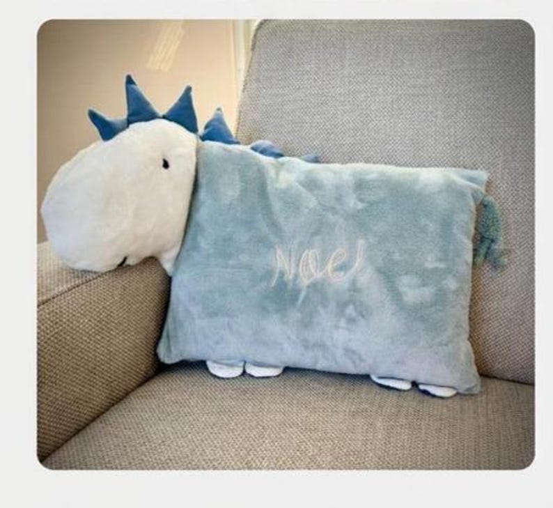 May include: A plush dinosaur-shaped pillow in white and light blue, with the name "Noel" embroidered on the pillow. The dinosaur has blue spikes and a fluffy white head. The pillow is resting on a gray upholstered chair.