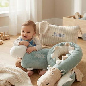 May include: A baby smiles while surrounded by plush toys. The toys include a blue and white dinosaur, a beige elephant pillow with the name "Mattia", and a green and white dinosaur-shaped neck pillow. A beige blanket is also present.