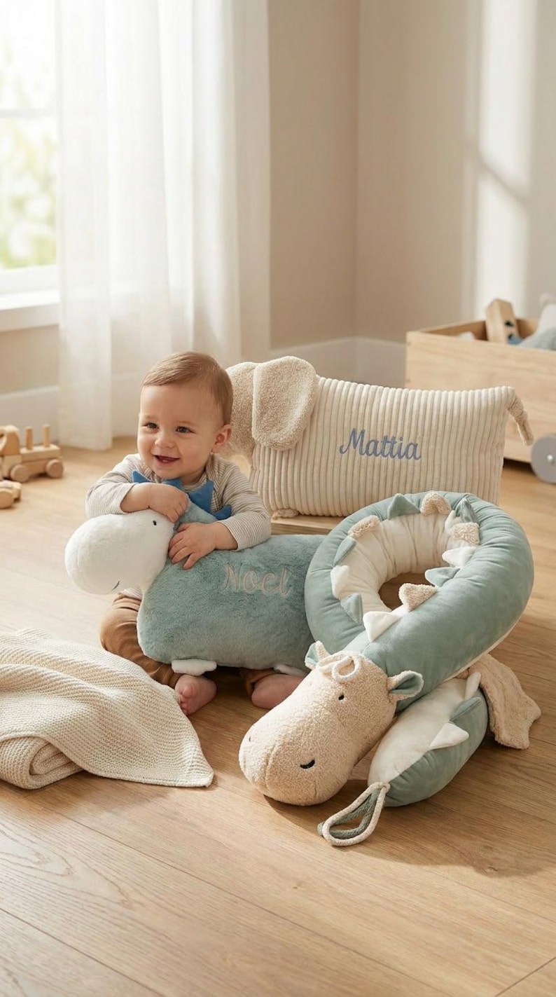 May include: A baby smiles while surrounded by plush toys. The toys include a blue and white dinosaur, a beige elephant pillow with the name "Mattia", and a green and white dinosaur-shaped neck pillow. A beige blanket is also present.