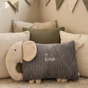 Peut inclure: Un coussin rectangulaire en velours c&ocirc;tel&eacute; gris fonc&eacute; en forme d'&eacute;l&eacute;phant avec une t&ecirc;te, des oreilles et des pieds en peluche beige. Le pr&eacute;nom Tino est brod&eacute; en &eacute;criture cursive blanche au centre du corps en velours c&ocirc;tel&eacute; gris. Le coussin est plac&eacute; devant plusieurs autres coussins d&eacute;coratifs aux tons neutres.