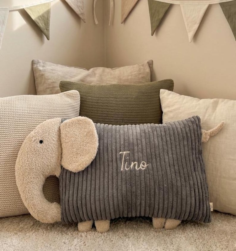 Peut inclure: Un coussin rectangulaire en velours c&ocirc;tel&eacute; gris fonc&eacute; en forme d'&eacute;l&eacute;phant avec une t&ecirc;te, des oreilles et des pieds en peluche beige. Le pr&eacute;nom Tino est brod&eacute; en &eacute;criture cursive blanche au centre du corps en velours c&ocirc;tel&eacute; gris. Le coussin est plac&eacute; devant plusieurs autres coussins d&eacute;coratifs aux tons neutres.