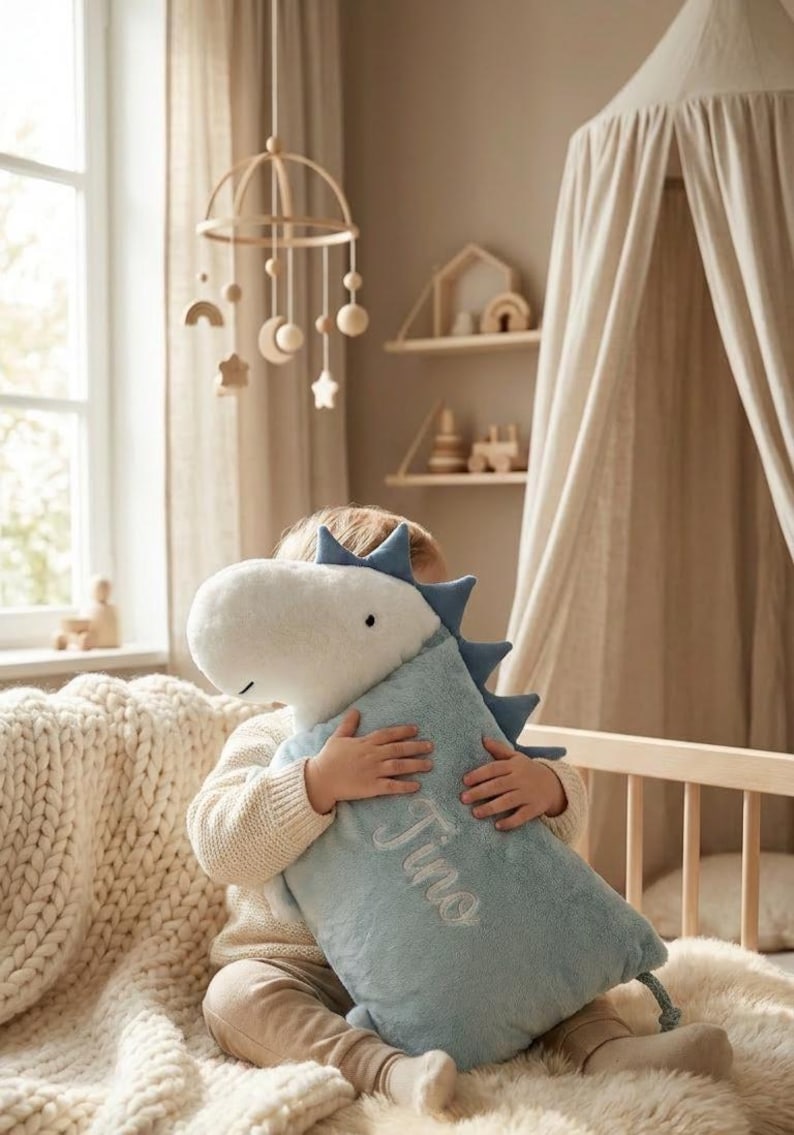 May include: A light blue dinosaur-shaped plush pillow with the name "Tino" embroidered on it. The pillow has a white head and blue spikes. A wooden mobile with moon and star shapes hangs in the background.