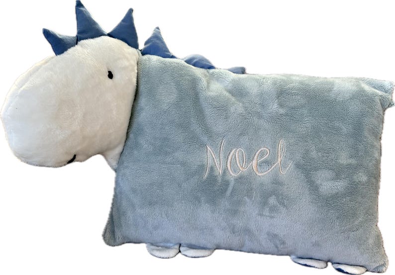 Könnte beinhalten: Ein hellblaues, plüschiges Kissen in Dinosaurierform mit dem Namen "Noel" in Schreibschrift gestickt. Der Dinosaurierkopf ist weiß mit blauen Akzenten und kleinen schwarzen Augen. Das Kissen hat eine weiche, flauschige Textur.