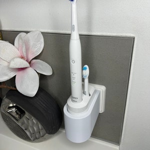 Könnte beinhalten: Eine weiße elektrische Zahnbürste und eine manuelle Zahnbürste sind an eine weiße, wandmontierte Ladestation angeschlossen. Die Ladestation hat einen runden Sockel und eine rechteckige Oberseite mit zwei Steckplätzen für die Zahnbürsten. Die Zahnbürsten sind beide weiß mit blauen Akzenten.