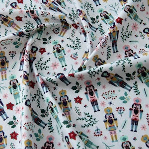 May include: White fabric with a repeating pattern of nutcracker soldiers in various colors, interspersed with sprigs of greenery, red poinsettias, and small pink flowers. The fabric is likely cotton or a similar material, suitable for quilting or crafting.