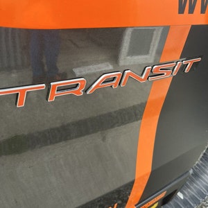 Könnte beinhalten: Nahaufnahme der Heckverkleidung eines Fahrzeugs mit dem Wort "TRANSIT" in silbernen und orangefarbenen Buchstaben. Die Verkleidung ist dunkelgrau mit orangefarbenen Streifen und Akzenten. Der obere Bereich ist orange.