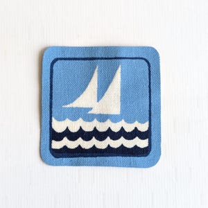 May include: Square, light blue fabric patch with a navy blue border. The patch features a design of two white sailboats on navy blue and white waves. The background is a lighter shade of blue.