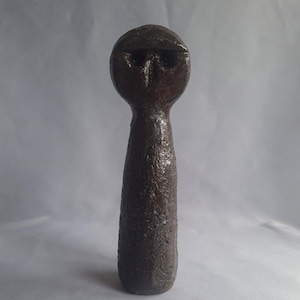 May include: A dark brown ceramic sculpture of a person with a rounded head and a long, slender body.