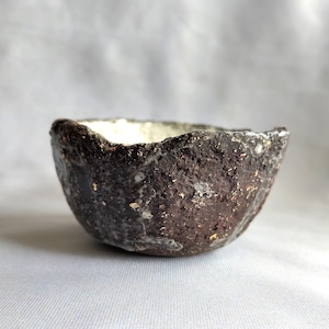 Peut inclure: Un petit bol en céramique brun foncé avec une surface rugueuse et texturée et un intérieur blanc. Le bol a un bord irrégulier, légèrement ondulé.