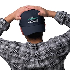 May include: A navy blue baseball cap featuring the text "2025 BELMONT at SARATOGA" in white and teal. The cap is held by a person wearing a black and white checked shirt.
