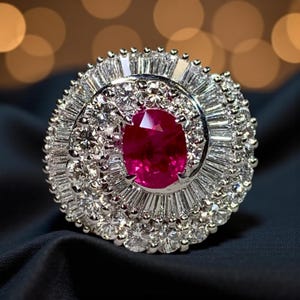 Puede incluir: Un anillo deslumbrante con un gran rubí ovalado en el centro, rodeado por un halo de diamantes claros y brillantes. Los diamantes están engastados en una banda de plata u oro blanco, creando una pieza de joyería lujosa y llamativa.
