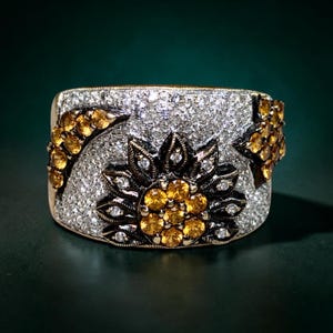 May include: A wide band ring featuring a central flower motif with yellow stones and dark metal petals. The background of the ring is covered in small clear stones, with additional yellow stone accents shaped like a crescent moon and a star.