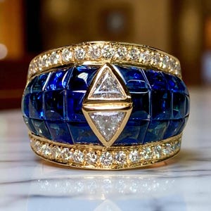 May include: A gold ring with a wide band featuring a central section of square-cut, deep blue gemstones. The ring is accented with a row of small diamonds and a gold triangle with two white diamonds.