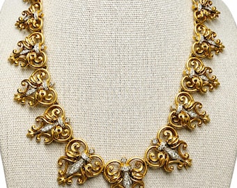 Könnte beinhalten: Eine goldfarbene Halskette mit einem verzierten Design. Die Halskette weist sich wiederholende, stilisierte Blumen- und Scrollwork-Elemente auf, die jeweils mit kleinen, funkelnden, klaren Steinen akzentuiert sind. Die Halskette wird auf einer neutralfarbenen Schaufensterpuppe präsentiert.