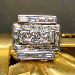May include: A silver ring with a large, round, center diamond surrounded by smaller diamonds. The ring has a geometric design with multiple rows of baguette-cut diamonds.