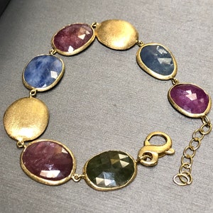 May include: A gold bracelet with a chain clasp features oval-shaped gemstones in shades of blue, green, and pink.