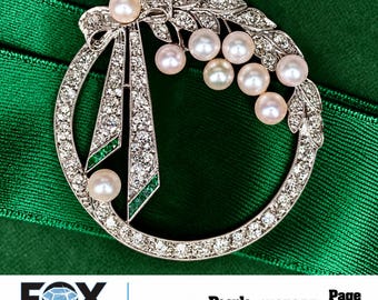 May include: A circular brooch featuring a silver-toned frame encrusted with clear stones, accented by a ribbon motif with green stones and multiple lustrous white pearls arranged in a cluster. The design includes a bow detail at the top.