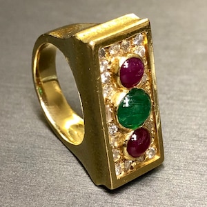 May include: A gold ring with a rectangular setting featuring three gemstones: a green emerald in the center, flanked by two red rubies. The setting is surrounded by smaller diamonds.