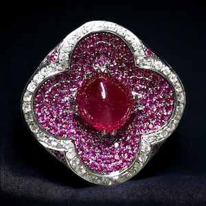 May include: A large, red ruby gemstone ring set in a white metal band. The ring is surrounded by a cluster of smaller red rubies and white diamonds.