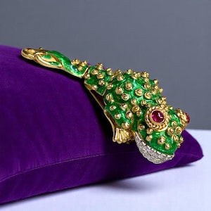 May include: A gold-toned frog brooch with green enamel, gold accents, and red gemstone eyes. The frog's mouth is lined with small, clear gemstones. The brooch is resting on a purple velvet surface.