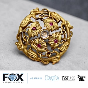 May include: A circular gold brooch featuring a central clear round gemstone surrounded by four gold leaves, each accented with a small red gemstone. The outer frame consists of a textured, vine-like pattern. Text at the bottom reads: FOX ESTATE JEWELRY AS SEEN IN People INSTORE Page Six.