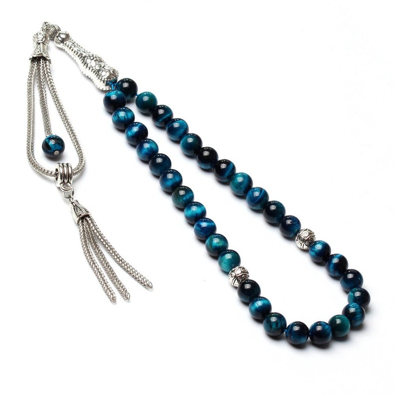 Puede incluir: Un rosario con cuentas de ojo de tigre azul. Las cuentas redondas presentan un patr&oacute;n arremolinado de azul y negro. Est&aacute;n ensartadas en una cadena plateada, con detalles plateados decorativos y una borla.