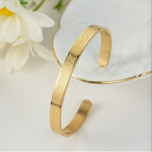 May include: A gold-colored cuff bracelet with engraved Arabic script. The open-ended bracelet is displayed on a white surface with a decorative floral element in the background. The bracelet has a polished, reflective surface.