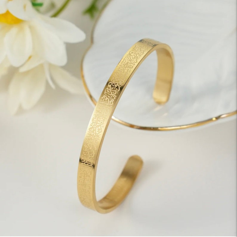 May include: A gold-colored cuff bracelet with engraved Arabic script. The open-ended bracelet is displayed on a white surface with a decorative floral element in the background. The bracelet has a polished, reflective surface.