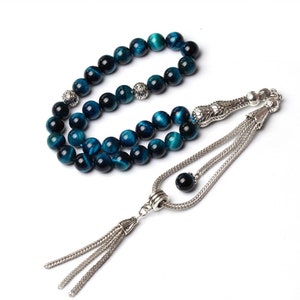Puede incluir: Un collar de cuentas de oraci&oacute;n con cuentas de ojo de tigre azul y detalles met&aacute;licos plateados. Las cuentas son una mezcla de azul oscuro y turquesa, con un acabado pulido. El collar tiene una borla decorativa.
