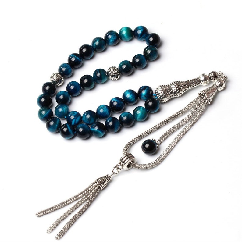 Puede incluir: Un collar de cuentas de oraci&oacute;n con cuentas de ojo de tigre azul y detalles met&aacute;licos plateados. Las cuentas son una mezcla de azul oscuro y turquesa, con un acabado pulido. El collar tiene una borla decorativa.