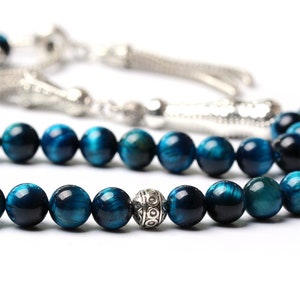 Puede incluir: Primer plano de un collar de cuentas de piedra preciosa ojo de tigre azul. Las cuentas redondas y pulidas tienen tonos azules y turquesas. Tambi&eacute;n se ve una cuenta y borla plateadas.