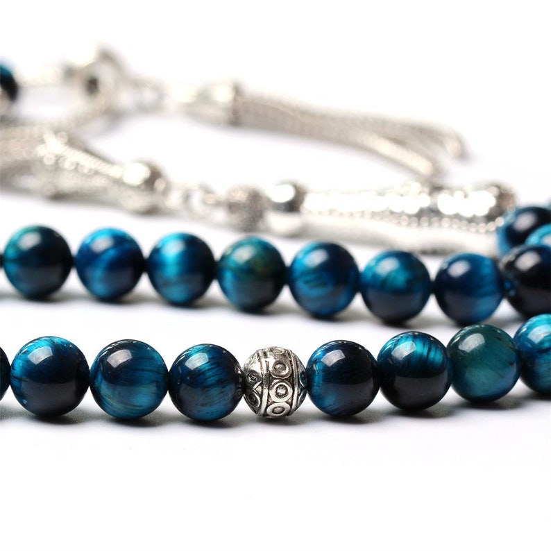 Puede incluir: Primer plano de un collar de cuentas de piedra preciosa ojo de tigre azul. Las cuentas redondas y pulidas tienen tonos azules y turquesas. Tambi&eacute;n se ve una cuenta y borla plateadas.