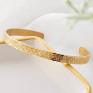May include: A gold-colored cuff bracelet with engraved Arabic script. The open-ended bracelet is resting on a gold-colored stick against a white background. The bracelet is a simple, elegant design.