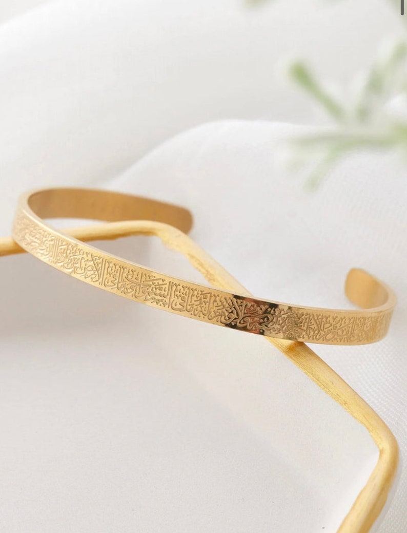 May include: A gold-colored cuff bracelet with engraved Arabic script. The open-ended bracelet is resting on a gold-colored stick against a white background. The bracelet is a simple, elegant design.