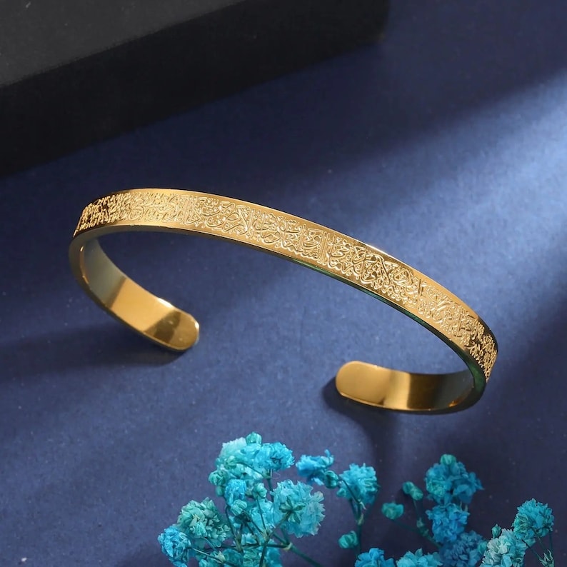 May include: A gold-colored cuff bracelet with intricate engraved detailing. The open-ended bracelet is curved and sits on a blue surface with blue flowers. The engraved design appears to be script.