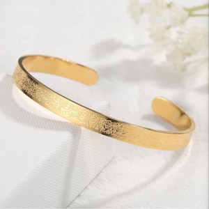 May include: A gold-colored open cuff bracelet. The bracelet features an engraved design along the top surface. The bracelet is displayed on a white surface with a soft, blurred background. The bracelet is a piece of jewelry.
