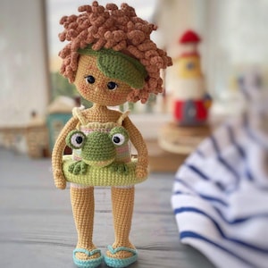 Pode incluir: Uma boneca de croch&ecirc; com cabelo castanho encaracolado usando um mai&ocirc; verde e segurando um sapo verde. A boneca est&aacute; usando chinelos azuis e est&aacute; em p&eacute; em uma superf&iacute;cie de madeira. A boneca est&aacute; usando uma faixa de cabe&ccedil;a verde.