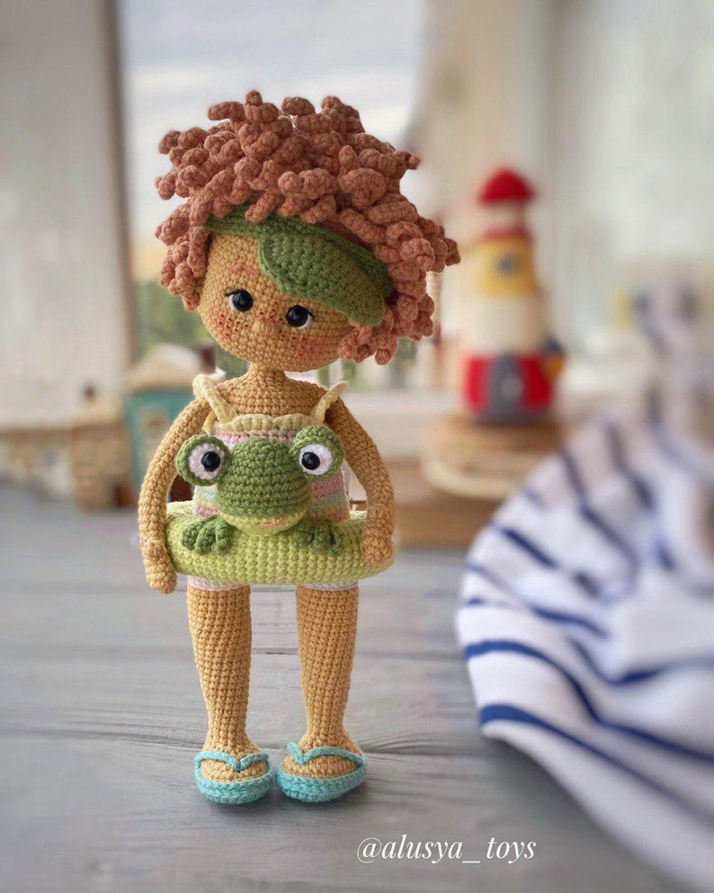 Pode incluir: Uma boneca de croch&ecirc; com cabelo castanho encaracolado usando um mai&ocirc; verde e segurando um sapo verde. A boneca est&aacute; usando chinelos azuis e est&aacute; em p&eacute; em uma superf&iacute;cie de madeira. A boneca est&aacute; usando uma faixa de cabe&ccedil;a verde.