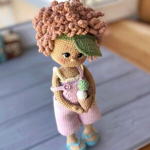 Pode incluir: Uma boneca de croch&ecirc; com cabelo castanho, vestindo uma roupa rosa e uma viseira verde, segurando um cone de sorvete. A boneca est&aacute; em p&eacute; sobre uma superf&iacute;cie de madeira azul claro. O texto "@alusiya_toys" &eacute; vis&iacute;vel no canto inferior direito da imagem.