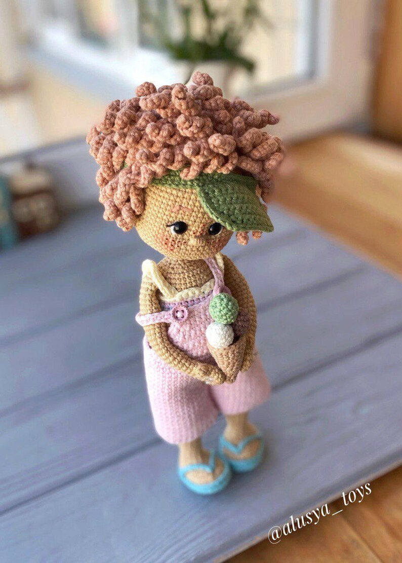 Pode incluir: Uma boneca de croch&ecirc; com cabelo castanho, vestindo uma roupa rosa e uma viseira verde, segurando um cone de sorvete. A boneca est&aacute; em p&eacute; sobre uma superf&iacute;cie de madeira azul claro. O texto "@alusiya_toys" &eacute; vis&iacute;vel no canto inferior direito da imagem.