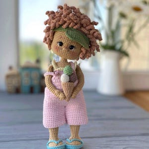 Pode incluir: Uma boneca de croch&ecirc; com cabelo castanho encaracolado, vestindo um macac&atilde;o rosa e uma faixa de cabe&ccedil;a verde, segurando um cone de sorvete. A boneca est&aacute; em p&eacute; sobre uma superf&iacute;cie de madeira. @alusya_toys
