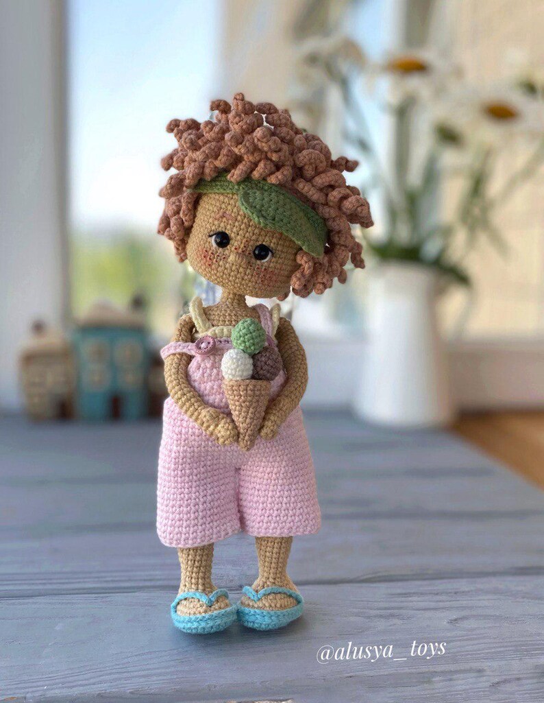Pode incluir: Uma boneca de croch&ecirc; com cabelo castanho encaracolado, vestindo um macac&atilde;o rosa e uma faixa de cabe&ccedil;a verde, segurando um cone de sorvete. A boneca est&aacute; em p&eacute; sobre uma superf&iacute;cie de madeira. @alusya_toys