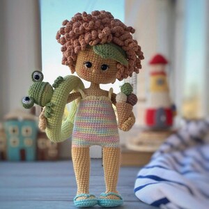Pode incluir: Uma boneca de croch&ecirc; com cabelo encaracolado castanho, vestindo um mai&ocirc; listrado e segurando um flutuador de sapo verde e um cone de sorvete. A boneca est&aacute; em p&eacute; sobre uma superf&iacute;cie de madeira com uma toalha listrada azul e branca no fundo.