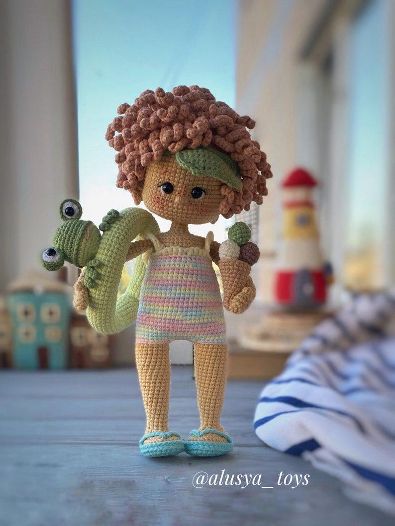 Pode incluir: Uma boneca de croch&ecirc; com cabelo encaracolado castanho, vestindo um mai&ocirc; listrado e segurando um flutuador de sapo verde e um cone de sorvete. A boneca est&aacute; em p&eacute; sobre uma superf&iacute;cie de madeira com uma toalha listrada azul e branca no fundo.