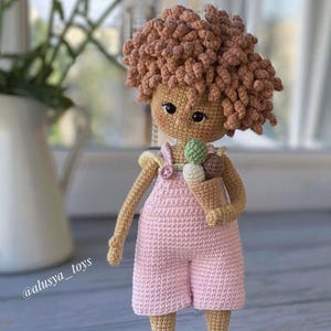 Pode incluir: Uma boneca de croch&ecirc; com cabelo castanho encaracolado, vestindo um vestido rosa e segurando um cone de sorvete. A boneca est&aacute; em p&eacute; sobre uma superf&iacute;cie de madeira. @alusiya_toys
