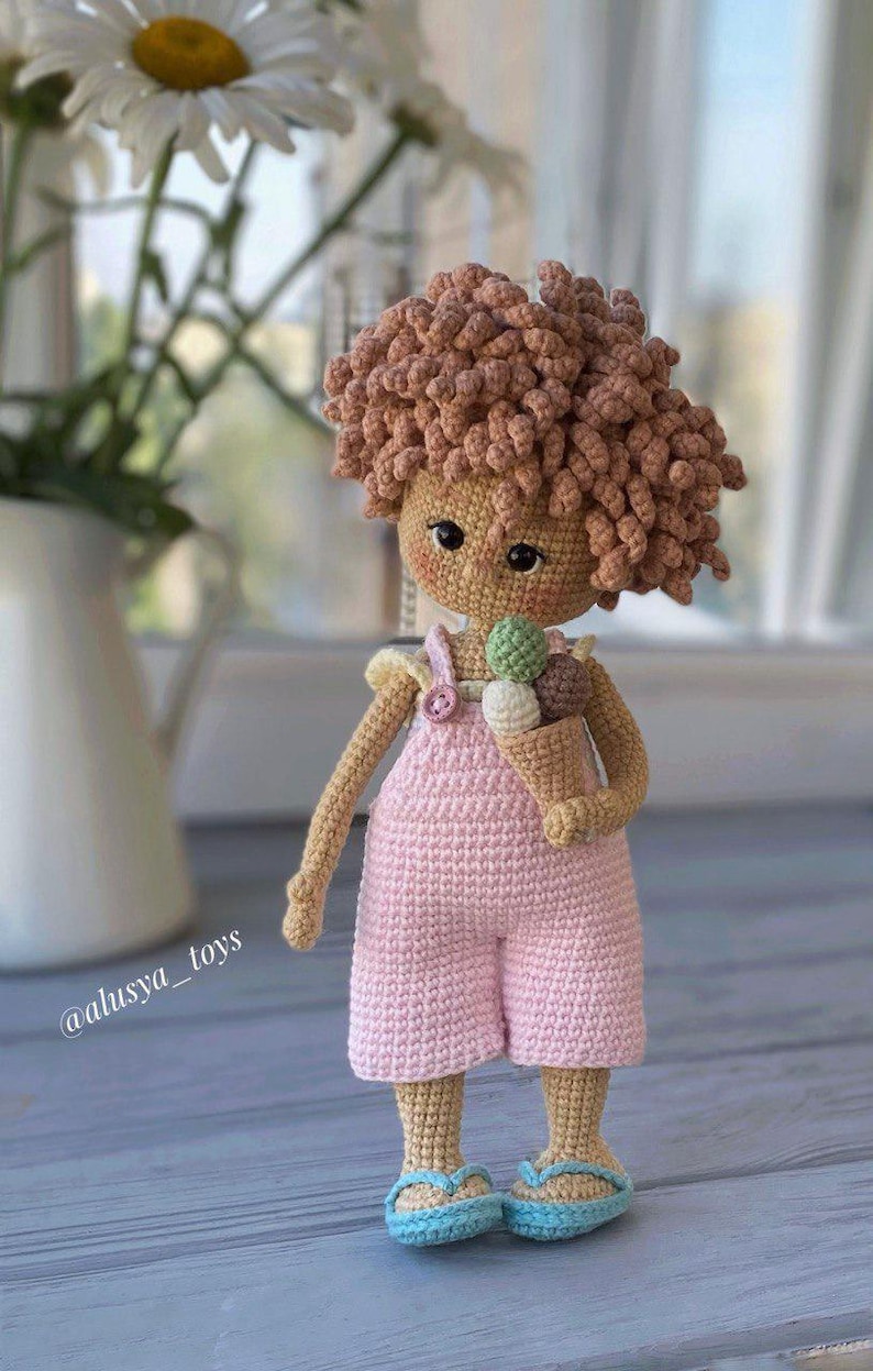 Pode incluir: Uma boneca de croch&ecirc; com cabelo castanho encaracolado, vestindo um vestido rosa e segurando um cone de sorvete. A boneca est&aacute; em p&eacute; sobre uma superf&iacute;cie de madeira. @alusiya_toys