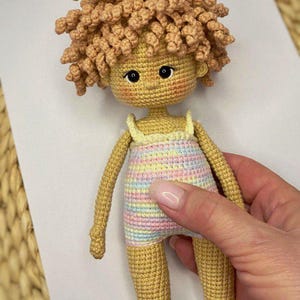 Pode incluir: Uma boneca de croch&ecirc; com cabelo encaracolado castanho claro, vestindo um vestido listrado pastel e sand&aacute;lias azuis. A boneca est&aacute; em p&eacute; sobre uma superf&iacute;cie branca.