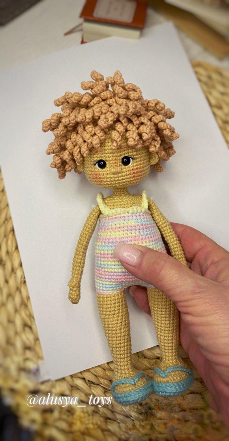 Pode incluir: Uma boneca de croch&ecirc; com cabelo encaracolado castanho claro, vestindo um vestido listrado pastel e sand&aacute;lias azuis. A boneca est&aacute; em p&eacute; sobre uma superf&iacute;cie branca.