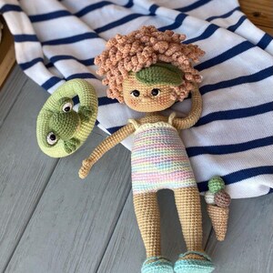 Pode incluir: Uma boneca de croch&ecirc; vestindo um mai&ocirc; listrado e segurando um cone de sorvete verde de croch&ecirc;. A boneca tem cabelo castanho encaracolado e usa sand&aacute;lias azuis. A boneca est&aacute; deitada em um cobertor listrado azul e branco.